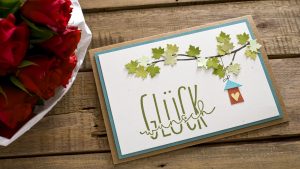 Ideen für Glückwünsche und Grußkarten: Glückwunsch | eine herzliche Papierszenerie in natürlichen Farben mit einem Ast mit Ahornblättern, einem Vogelhäuschen mit Herz und einem handgelettertem Schriftzug