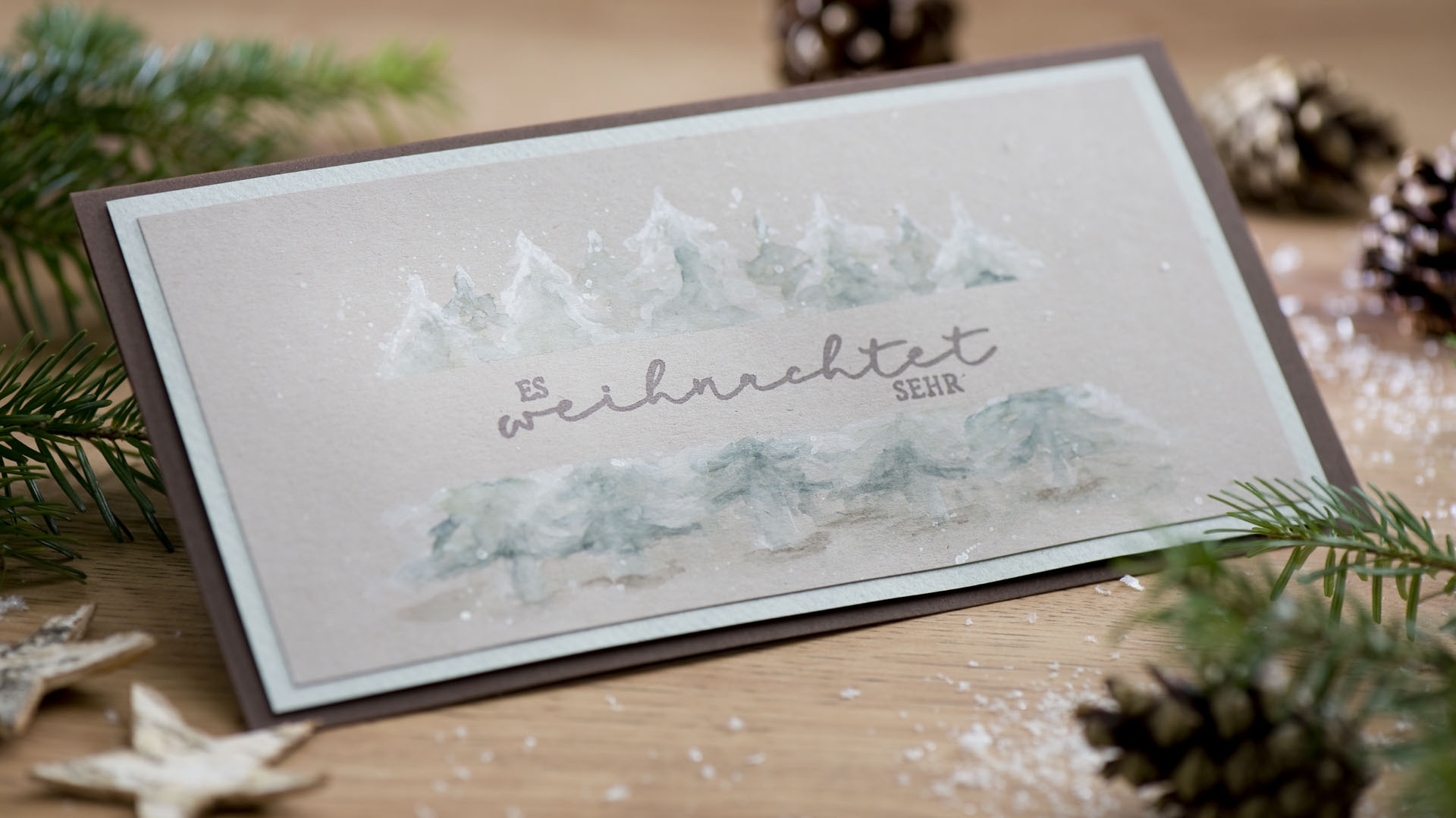 Ideen für die Weihnachtpost: Oh, Tannenbäume! Es weihnachtet sehr. | Handgemachte Weihnachtskarte im Querformat in natürlichen Farben mit einer Kulisse aus aquarellierten Tannenbäumen und Schneeflocken mit einem gestempelten Schriftzug in der Mitte.